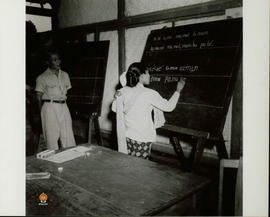 Seorang ibu menggendong anaknya sedang belajar menulis di depan kelas dan didampingi oleh seorang...