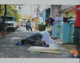 Dua orang sedang melakukan sholat di jalan dan di jaga oleh seorang warga.